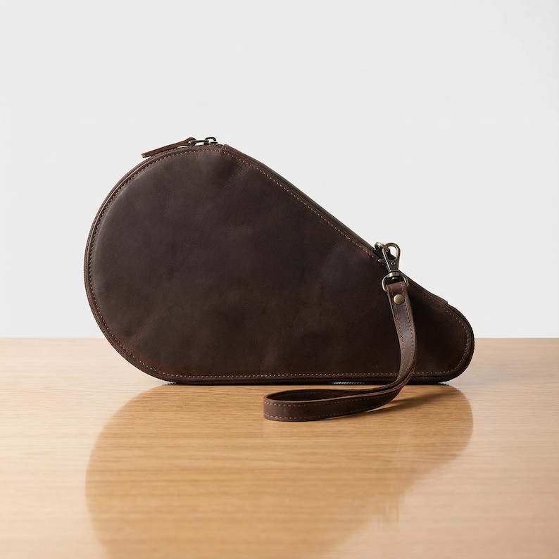 May include: A brown leather clutch with a rounded top and a zipper closure. The clutch has a wrist strap attached with a metal clasp. The bag is sitting on a wooden surface.
