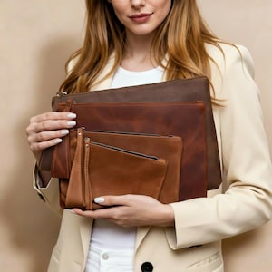 May include: A collection of brown leather pouches in varying sizes. The pouches have a zipper closure and a wrist strap on the smallest pouch. The leather has a distressed appearance, with shades ranging from dark brown to tan.