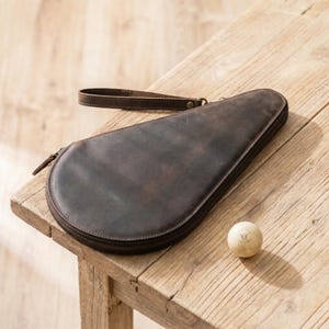 May include: A brown leather table tennis paddle case with a wrist strap and a zipper closure. A ping pong ball with a smiling face is next to the case. The case is on a wooden table.