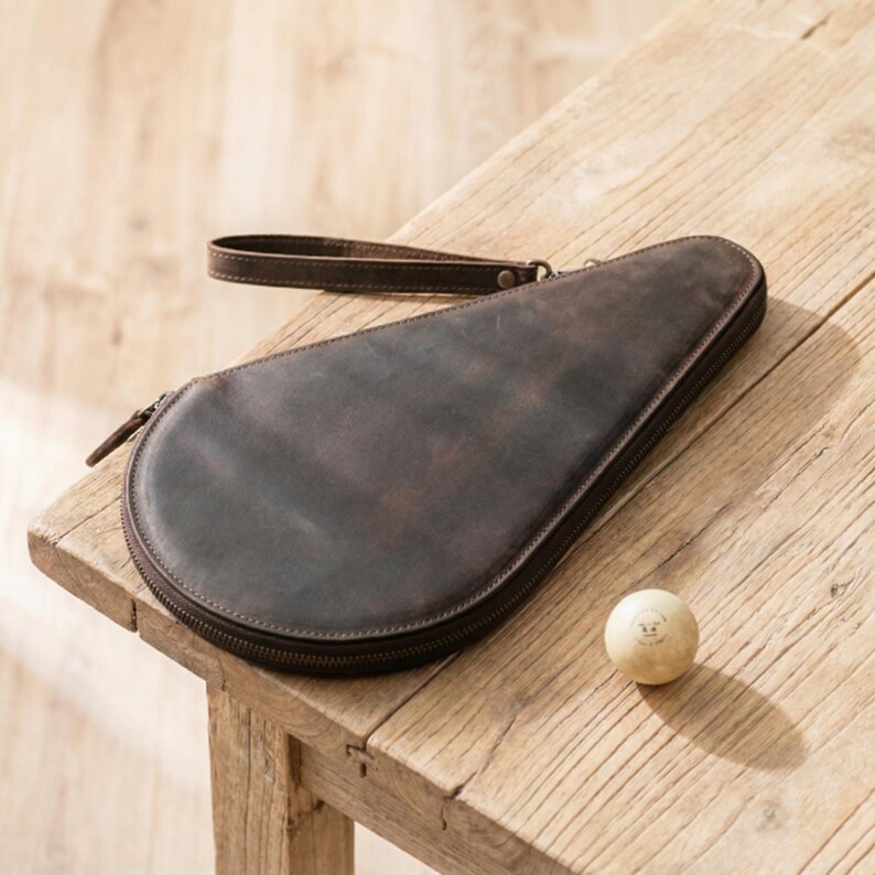 May include: A brown leather table tennis paddle case with a wrist strap and a zipper closure. A ping pong ball with a smiling face is next to the case. The case is on a wooden table.