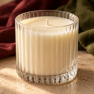 May include: A cylindrical candle in a clear, vertically ribbed glass container. The wax is off-white and opaque, featuring a single centered cotton wick. The glass vessel has a thick base and distinct vertical fluting around the exterior.