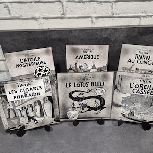 Könnte beinhalten: Eine Sammlung von Tintin-Comics in Schwarzweiß, präsentiert auf kleinen schwarzen Ständern. Die Cover zeigen verschiedene Titel, darunter "L'Étoile Mystérieuse", "Amérique", "Au Congo", "Les Cigares du Pharaon", "Le Lotus Bleu" und "L'Oreille Cassée".