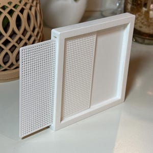 Puede incluir: Un marco rectangular blanco con un panel deslizante que presenta una rejilla de pequeñas perforaciones circulares. El marco tiene un borde liso y una sección empotrada que sostiene la pieza deslizante perforada, creando un diseño funcional.