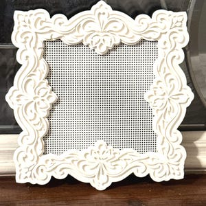 May include: A square white frame featuring an intricate, laser-cut filigree border with symmetrical floral-inspired scrollwork patterns. The center of the frame is filled with a fine white mesh screen that has a uniform grid of small circular holes.