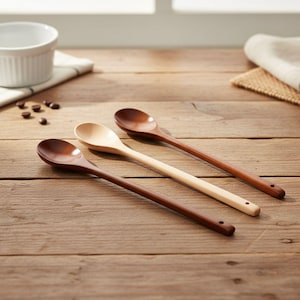 Può includere: Tre cucchiai di legno in varie tonalità di marrone e legno naturale sono disposti diagonalmente su una superficie di legno. I cucchiai hanno manici lunghi e ciotole arrotondate, con un piccolo foro all'estremità di ogni manico. Una ciotola bianca e chicchi di caffè sullo sfondo.