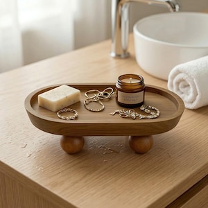 Puede incluir: Una bandeja ovalada de madera con bordes redondeados, sostenida por dos pies esféricos. La bandeja contiene una pastilla de jabón, joyas y una pequeña vela con la etiqueta "Sandalwood & Sea Salt". Los artículos están dispuestos sobre una superficie de madera clara.