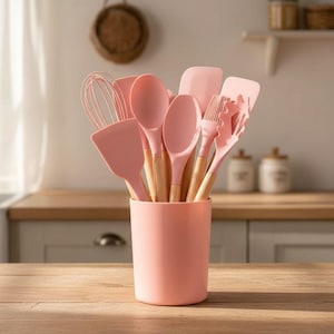 Peut inclure: Un ensemble d'ustensiles de cuisine roses, comprenant un fouet, des spatules et des cuillères, avec des manches en bois, est disposé dans un récipient cylindrique rose assorti. L'ensemble est présenté sur une surface en bois, suggérant une cuisine.