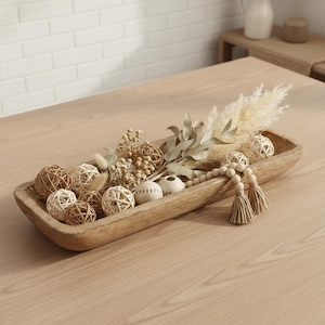Può includere: Una ciotola decorativa in legno piena di sfere decorative, fiori secchi e una ghirlanda di nappe di perline. La ciotola è marrone chiaro e poggia su un tavolo di legno. Le sfere sono in varie tonalità di marrone e bianco.