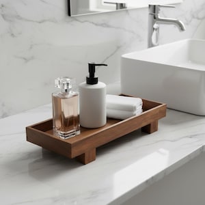 May include: A wooden bathroom tray holds a perfume bottle, a white soap dispenser with a black pump, and folded white towels. The tray sits on a white marble countertop near a white sink and faucet.