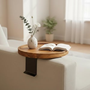 May include: A round wooden sofa arm tray with a black metal clip attachment. The tray holds a small white ceramic vase containing a sprig of eucalyptus and a white flower, alongside an open book with white pages resting on the wood surface.