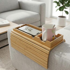 May include: A bamboo sofa arm tray with a built-in phone holder and a cup holder. A pink mug with a white handle and a spoon rests in the cup holder. A smartphone is in the phone holder. The tray is on a gray sofa arm.