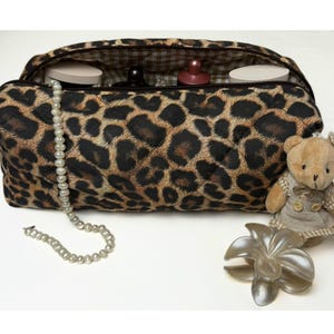 Könnte beinhalten: Eine Kosmetiktasche mit Leopardenmuster und Steppdesign, geöffnet, um Schönheitsprodukte zu zeigen. Eine Perlenkette, ein kleiner Teddybär und eine dekorative Blume liegen neben der Tasche. Die Tasche ist braun und schwarz.