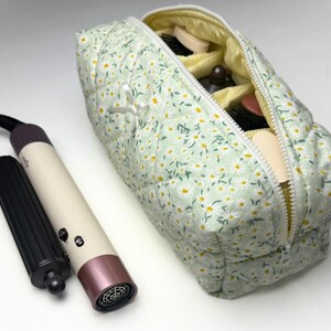 Könnte beinhalten: Eine hellgrüne, gesteppte Kosmetiktasche mit Gänseblümchenmuster, teilweise geöffnet, um Make-up-Artikel zu zeigen. Ein weißer und roségoldener Haartrockner und ein schwarzer Aufsatz liegen neben der Tasche.
