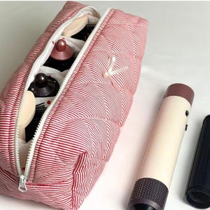 May include: A red and white striped quilted cosmetic bag with a white zipper, open to reveal makeup items. A beige and brown hair styling tool and a black attachment are next to the bag.