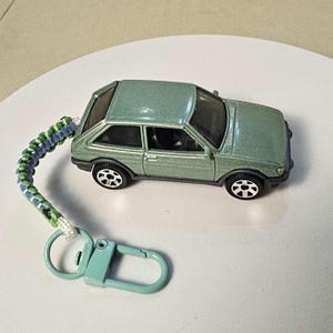 May include: A metallic sage green die-cast hatchback toy car keychain featuring a braided paracord lanyard in shades of green, blue, and white, attached to a light green metal lobster clasp and a circular split ring for keys.