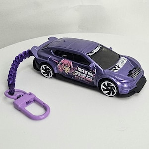 May include: A purple die-cast toy car with a keychain. The car features black wheels and a graphic of an anime character and text. The keychain is made of braided cord and a purple clasp.