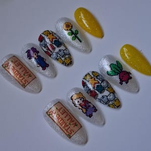 May include: A set of ten almond-shaped press-on nails with a white glitter base. Designs include the Stardew Valley logo, pixelated character figures, a sunflower, a radish, and a pattern of colorful pixelated gems on a white background.