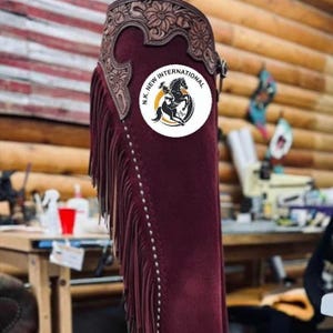 May include: Burgundy leather chaps with decorative floral tooling at the top and fringe along the side. The chaps feature a white circular logo with a horse and the text "N.K. NEW INTERNATIONAL". The chaps are designed for horseback riding.