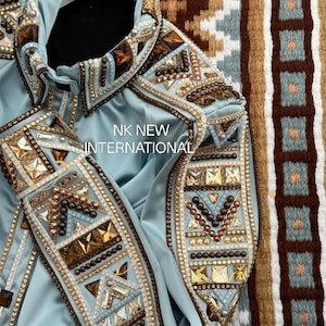 Könnte beinhalten: Hellblaue Jacke mit aufwendigen Verzierungen. Geometrische Muster mit braunen, goldenen und klaren Kristallen. Der Text "NK NEW INTERNATIONAL" ist auf der Vorderseite zu sehen.