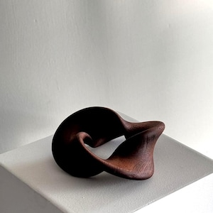 May include: A dark brown wooden sculpture with an abstract, looped design. The sculpture is displayed on a white surface, highlighting its smooth curves and organic form. The handcrafted piece has visible wood grain, suggesting a natural aesthetic.
