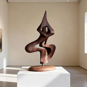 May include: A dark brown wooden sculpture with an abstract design. The sculpture has a tall, pointed top and a flowing, curved shape. It is mounted on a round, wooden base and displayed on a white pedestal in a gallery setting.