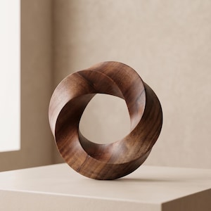 Puede incluir: Una escultura de madera con un diseño de bucle continuo e entrelazado. La escultura está hecha de madera marrón oscuro y se encuentra sobre una superficie beige claro. Es una obra de arte moderna y abstracta.