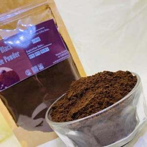 Può includere: Una ciotola di vetro trasparente riempita di polvere di caffè marrone scuro si trova davanti a un sacchetto di carta marrone. L'etichetta del sacchetto riporta il testo Black Coffee Powder, un peso di 100g, scadenza 09 09 2024 e icone non-OGM.