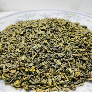 Può includere: Primo piano di un mucchio di foglie di tè verde essiccate. Le foglie, di colore verde chiaro e scuro, hanno un aspetto leggermente arricciato. Il tè è su un piatto bianco con un motivo floreale.