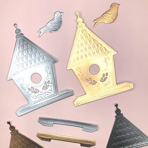 May include: A collection of metal craft dies featuring two birdhouse shapes in silver and gold, two small bird silhouettes in silver and gold, and two rectangular base strips in silver and gold, all with embossed roof and floral details.
