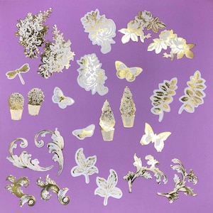 May include: A collection of gold and white decorative stickers on a purple background. The stickers include floral designs, butterflies, leaves, and decorative flourishes. The stickers are in various shapes and sizes, with a metallic gold finish.