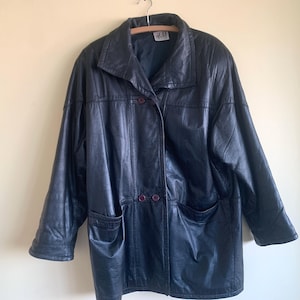 May include: A vintage navy blue leather jacket. It features a collared neckline, two front pockets, and button fastenings. The jacket is suspended on a wooden hanger. The leather has a slightly worn appearance, giving it a classic, timeless look.