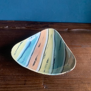 Op de afbeelding: Een kleurrijke, driehoekige keramische schaal met een gestreept patroon in geel, perzik, blauw en groen, geaccentueerd met zwarte lijnen. De schaal heeft een gouden rand en staat op een houten oppervlak.