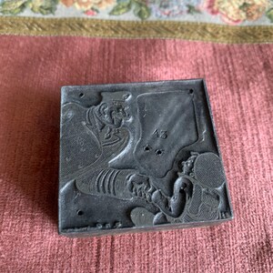 May include: A dark gray, rectangular printing block with detailed relief carvings of figures and the number "43". The block has a worn appearance, suggesting age and use. The background is a pink fabric with floral trim.
