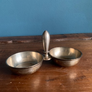 Può includere: Un piatto doppio argentato con manico centrale. Le due ciotole ovali sono affiancate, collegate da un piccolo piedistallo. Il manico è allungato e bulboso. Il pezzo è appoggiato su una superficie di legno.