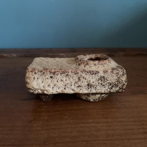 May include: A small, handmade ceramic piece with a textured, speckled surface. The object is rectangular with a circular indentation on top and rests on two small, rounded feet. The color is a mix of beige and brown.