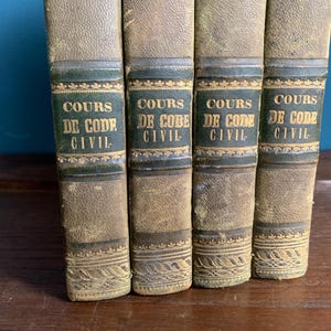 Puede incluir: Cuatro libros antiguos con cubiertas de cuero envejecido. Las lomos son de color verde oscuro con letras doradas que dicen "COURS DE CODE CIVIL". Los libros están colocados uno al lado del otro, sugiriendo una colección de textos legales.