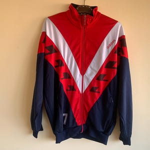 Op de afbeelding: Een vintage Adidas trainingsjack in rood, wit en marineblauw. De jas heeft een volledige ritssluiting, een hoge kraag en het Adidas-logo op de borst. Het ontwerp omvat een chevronpatroon en het nummer 7 op de mouw.