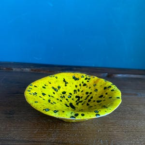 Op de afbeelding: Een kleine, ronde, gele glazen kom met zwarte spikkels. De kom heeft een onregelmatige vorm en staat op een houten oppervlak. De achtergrond is effen blauw.