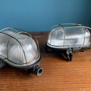 Op de afbeelding: Twee buitenlampen in vintage stijl. Elke lamp heeft een heldere, geribbelde glazen koepel in een metalen kooi, gemonteerd op een zwarte basis. De lampen zijn ontworpen voor wandmontage en zijn geschikt voor buitengebruik.