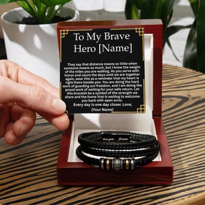 May include: A black braided leather bracelet with silver and gold accents, presented in a dark wooden box. The box lid features a heartfelt message, including the words "To My Brave Hero" and "love you forever" on the bracelet clasp.
