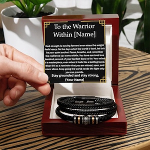 May include: A black braided leather bracelet with silver and gold accents, presented in a wooden box. The box lid features the text "To the Warrior Within [Name]" and a motivational message. The bracelet is inscribed with "love you forever".