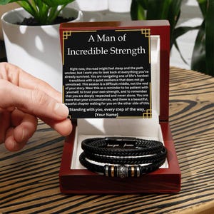 May include: A black leather bracelet with silver and gold accents is displayed inside a red wooden box. The box contains a card with the text "A Man of Incredible Strength" and a message. The bracelet has the words "love you forever" engraved on it.