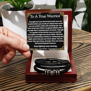 May include: A black braided leather bracelet with silver and gold accents, presented in a dark wooden box. The box contains a card with the text "To A True Warrior" and the words "love you forever" on the bracelet.