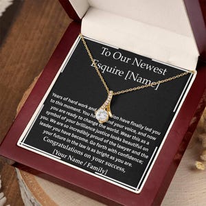 May include: A gold-colored necklace with a pendant featuring a large, clear gemstone and smaller accent stones. The necklace is presented in a wooden gift box with a black card that reads "To Our Newest Esquire [Name]" and a congratulatory message.