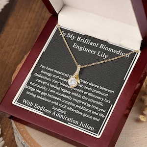 May include: A gold-colored pendant necklace with a clear, round gemstone in a gift box. The box has a black background with white text that reads "To My Brilliant Biomedical Engineer Lily" and a message from Julian.