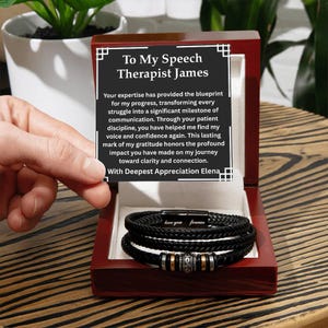 May include: A black leather bracelet with silver and gold accents, presented in a wooden gift box. The box contains a message that reads "To My Speech Therapist James" and "love you forever" on the bracelet.