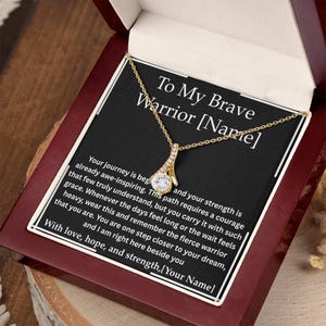 May include: A gold-colored pendant necklace with a clear, round gemstone in a gift box. The necklace is displayed on a black card with the text "To My Brave Warrior [Name]" and a message of encouragement.