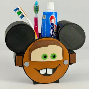 May include: A novelty toothbrush holder in the shape of a cartoon car character, with black ears, a brown face, and green eyes. It contains two toothbrushes and a tube of Crest Kids toothpaste.