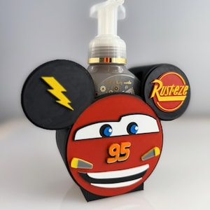 May include: A red and black soap dispenser holder shaped like Lightning McQueen from the Cars movie. The holder has black ears with a yellow lightning bolt and the Rusteze logo. The number 95 is visible on the front.