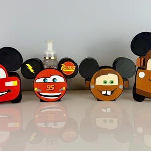 May include: A selection of car-shaped decorations with Mickey Mouse ears. The cars are red, orange, and brown, with smiling faces and names like "Luca" and "Kaden". A clear bottle with a pump is in the centre.